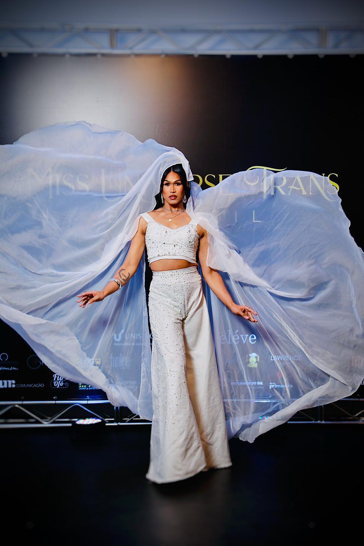 Miss Universe Trans Brasil em um vestido branco deslumbrante com um véu esvoaçante, em frente ao logo do evento.