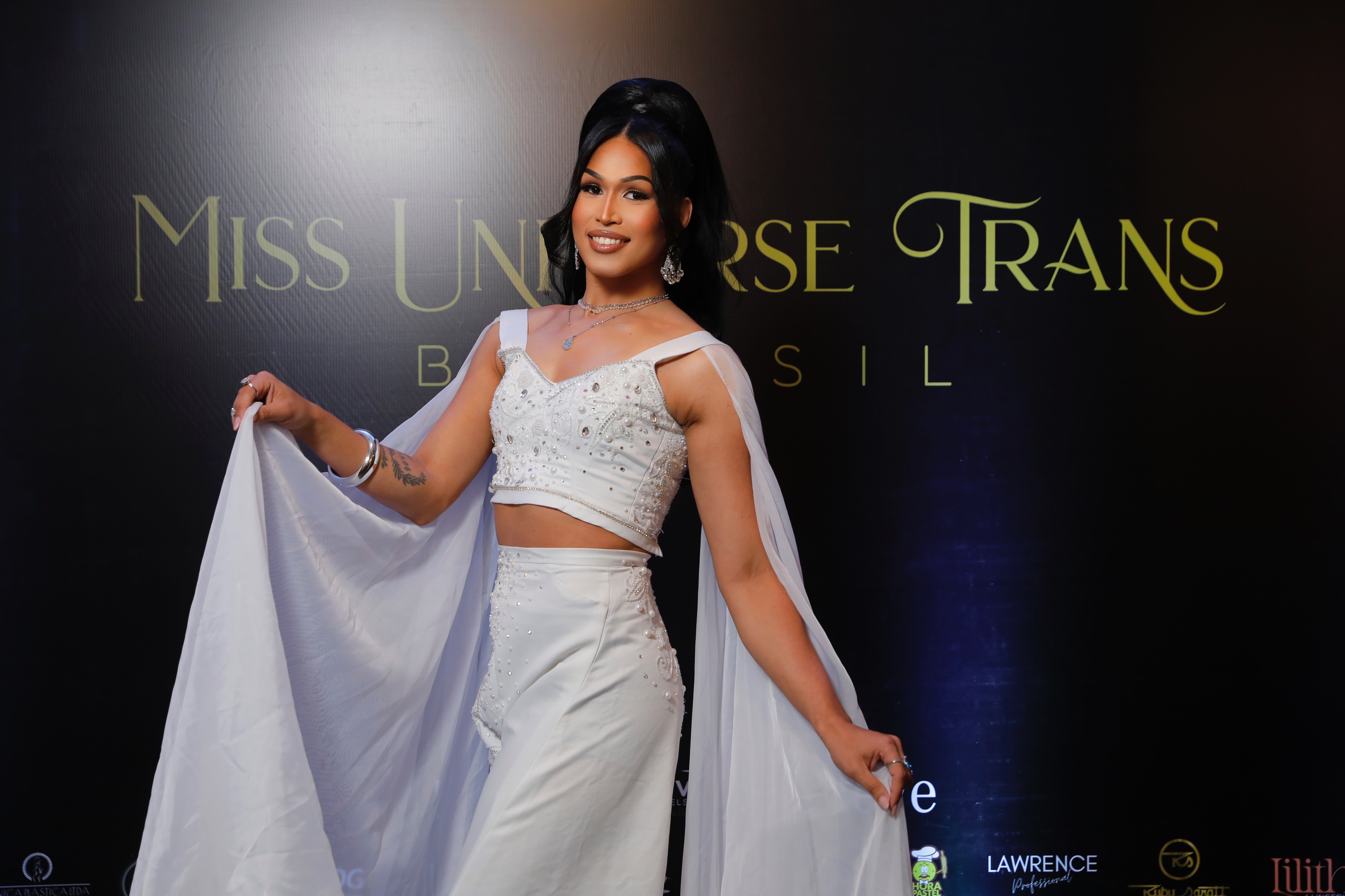 Miss Universe Trans Brasil em um conjunto branco bordado, sorrindo, com o logo do evento ao fundo.